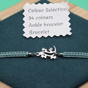 May include: A teal macrame bracelet with a silver lizard charm. The bracelet is displayed on a dark teal hexagonal fabric. A white tag above the bracelet reads "Colour Selection: 34 colours Ankle bracelet Bracelet."