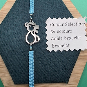 May include: A light blue macrame bracelet with a silver monkey charm. The bracelet is displayed on a dark teal surface. Text on a white tag reads "Colour Selection: 34 colours Ankle bracelet Bracelet."