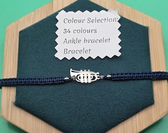 May include: A navy blue macrame bracelet with a silver trumpet charm. The bracelet is displayed on a dark green fabric, set on a wooden hexagonal base. A white tag indicates color options and the item's name.