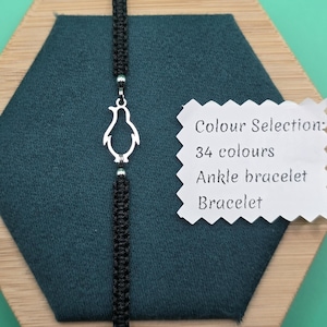 May include: Black woven bracelet featuring a silver penguin charm. The bracelet is presented on a dark teal surface. A white tag states "Colour Selection: 34 colours, Ankle bracelet, Bracelet."