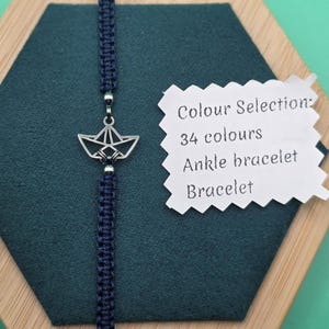 Op de afbeelding: Marineblauwe macramé enkelband met een zilverkleurige origami boot bedel. De armband wordt getoond op een donkergroen oppervlak. De tekst op het label luidt "Colour Selection: 34 colours Ankle bracelet Bracelet."