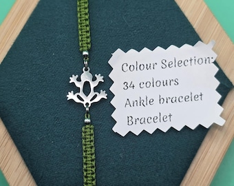 Könnte beinhalten: Ein grünes geflochtenes Armband mit einem silbernen Frosch-Anhänger. Das Armband wird auf einer dunkelgrünen Oberfläche mit einem Holzrahmen präsentiert. Ein weißes Etikett lautet "Colour Selection: 34 colours, Fußkettchen, Armband."