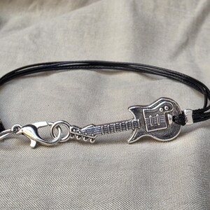May include: A black cord bracelet featuring a silver-toned electric guitar charm as the central focal point. The bracelet is secured with a silver-toned lobster claw clasp and a jump ring, connecting the guitar charm to the multi-strand black cord.