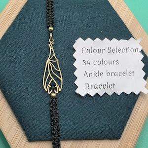 Può includere: Braccialetto alla caviglia in macramè nero con un ciondolo a forma di pipistrello color oro. Il ciondolo è una silhouette di pipistrello stilizzata. L'immagine include anche un'etichetta che dice "Colour Selection: 34 colours Ankle bracelet Bracelet."