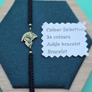 May include: Black macrame bracelet with a gold-colored Spartan helmet charm. The bracelet is displayed on a dark green surface with a white tag that reads "Colour Selection: 34 colours Ankle bracelet Bracelet."