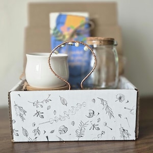 May include: A gift box with a white ceramic cup, a clear glass jar with a cork lid, and a copper-colored headband with small white beads. The box is decorated with botanical illustrations.
