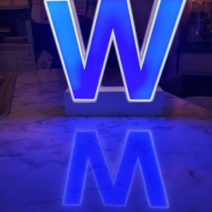 May include: A large, illuminated letter "W" in bright blue, with a white border, casting a glowing reflection. The letter sits on a white base, creating a striking visual effect. The background is blurred, focusing attention on the bold, modern design.