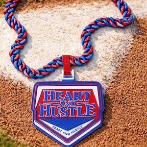 May include: A baseball-themed necklace with a blue, red, and purple braided chain. The pendant is a blue home plate shape with the words "HEART AND HUSTLE" in red and white. The necklace rests on a baseball field.