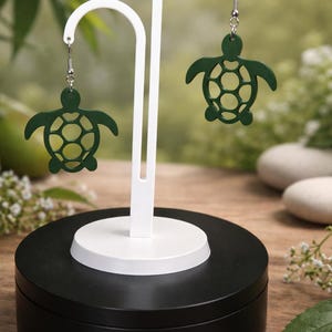 May include: Dark green turtle-shaped earrings suspended from a white display stand. The earrings feature a cut-out design and are presented on a black and white base. The background includes greenery and stones.