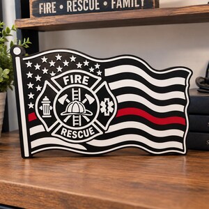 Peut inclure: Une enseigne rectangulaire en forme de drapeau ondulant avec un motif noir et blanc présentant un emblème central Fire Rescue. L'emblème comprend un casque, des haches et une borne d'incendie. Une bande rouge horizontale traverse le drapeau.