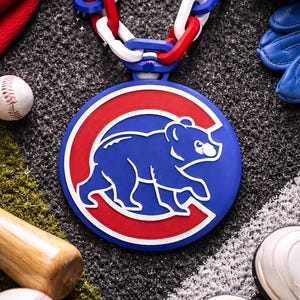May include: A blue, red, and white Chicago Cubs pendant necklace with a chain. The pendant features the team's logo, a blue bear inside a red circle. Baseball equipment, including a bat, ball, and glove, surrounds the necklace.