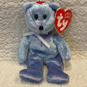 Può includere: Un orsacchiotto di peluche blu con un nastro bianco e un'etichetta rossa a forma di cuore. L'orso ha occhi neri e un naso nero. L'etichetta dice "Jingle Beanies" e "ty".
