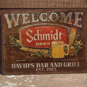 May include: A vintage-style metal sign with a rusty brown background. The sign says "Welcome" in large, white letters. It features a red and white logo for Schmidt Beer with the text "Proudly Serving" and "The Brew That Grew With The Great Northwest". Below the logo is the text "David's Bar and Grill Est. 1983".