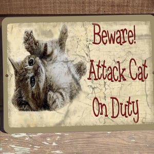 May include: A beige metal sign with a brown tabby cat curled up on it. The sign reads "Beware! Attack Cat On Duty".