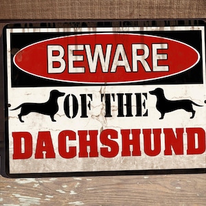 May include: A red, black, and white metal sign with the text "Beware of the Dachshund". The sign has a distressed look and features two black silhouettes of Dachshunds.