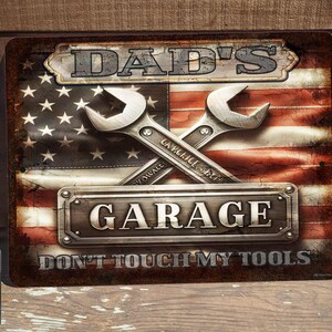 May include: A red, white, and blue American flag with two crossed wrenches. The wrenches are silver and have the words "Garage" and "Don't Touch My Tools" written on them. The flag has the words "Dad's Garage" written on it.