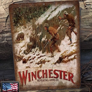 May include: Vintage metal sign featuring a hunting scene with two figures in a snowy forest. The sign has a distressed, rusty look and features the text "Winchester Repeating Arms Co."