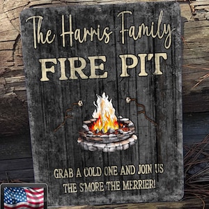 May include: A rectangular wooden sign with the text "The Harris Family FIRE PIT." A graphic of a campfire with flames and marshmallows on sticks is in the center. The text "GRAB A COLD ONE AND JOIN US THE S'MORE THE MERRIER!" is below. The sign has a "MADE IN U.S.A PRODUCT" logo.