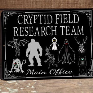 May include: Black and white sign with the text "Cryptid Field Research Team" and the words "Main Office" at the bottom. The sign features illustrations of various cryptids, including a Bigfoot, a Mothman, a Jersey Devil, a creature from the Black Lagoon, and a ghost.