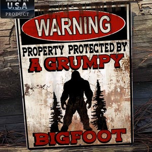 May include: A warning sign with a distressed, rusty metal look. The sign reads "Warning Property Protected by A Grumpy Bigfoot". The sign features a silhouette of Bigfoot standing between two pine trees.