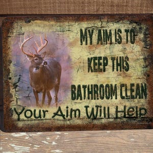 May include: Rustic metal sign with a distressed background featuring a deer and the text "My aim is to keep this bathroom clean. Your aim will help."