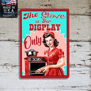 May include: A vintage-style metal sign with a red frame. The sign features a retro illustration of a woman in a red dress and the text "The Stove is For DISPLAY Only." An American flag and "Made in U.S.A. PRODUCT" are in the upper left corner.