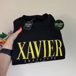 May include: A black crewneck sweatshirt featuring the yellow embroidered text XAVIER INSTITUTE EST 1963. The garment includes a black tag with the white text MISCHIEF & MAGIC and a circular green logo with the text MISCHIEF & MAGIC.
