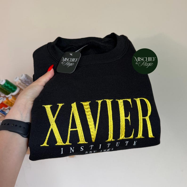 May include: A black crewneck sweatshirt featuring the yellow embroidered text XAVIER INSTITUTE EST 1963. The garment includes a black tag with the white text MISCHIEF & MAGIC and a circular green logo with the text MISCHIEF & MAGIC.