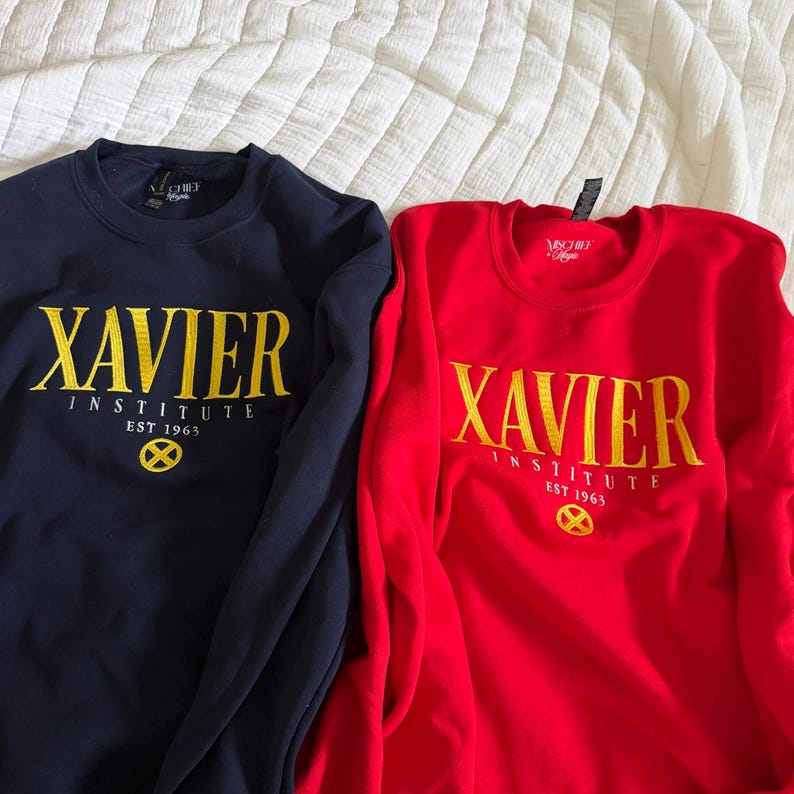 May include: A navy blue sweatshirt and a red sweatshirt, each featuring yellow embroidered text that reads XAVIER INSTITUTE EST 1963 above a yellow circular X-Men logo. Both garments have long sleeves, a crew neckline, and a ribbed collar.