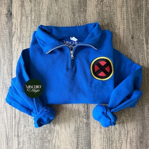 Puede incluir: Una sudadera azul brillante con cremallera de un cuarto de largo en metal plateado y un parche circular en el pecho. El parche muestra una X roja sobre un fondo negro, rodeada por un borde amarillo. La etiqueta interior dice Mischief & Magic.