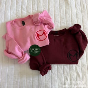 Op de afbeelding: Twee sweaters met ronde hals, één lichtroze en één donkerbordeaux, elk met een cirkelvormig geborduurd logo met een gestileerde letter M op de linkerborst. Een ronde groene sticker met de tekst MISCHIEF & MAGIC is ertussen geplaatst.
