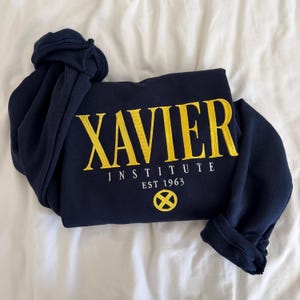 Puede incluir: Una sudadera azul marino de manga larga con texto bordado en amarillo que dice XAVIER INSTITUTE EST 1963. Debajo del texto hay un emblema circular amarillo con un diseño de X. La prenda tiene cuello redondo y puños acanalados.