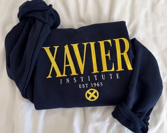May include: A navy blue long-sleeve sweatshirt featuring yellow embroidered text that reads XAVIER INSTITUTE EST 1963. Below the text is a circular yellow emblem with an X design. The garment has a crew neckline and ribbed cuffs.