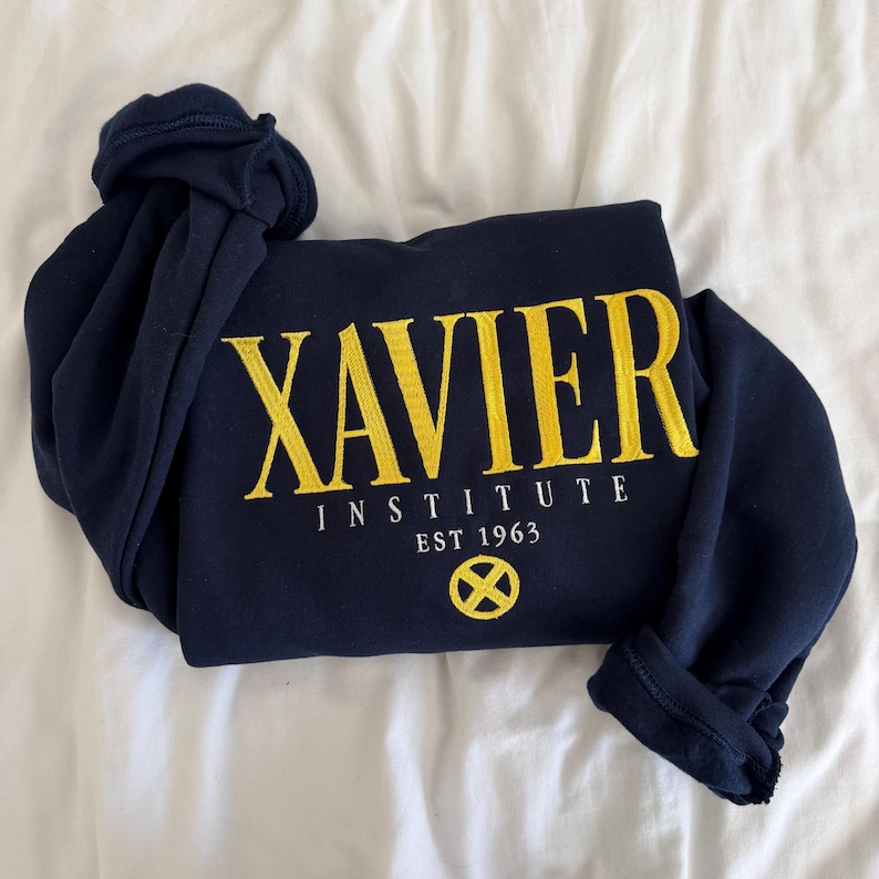 May include: A navy blue long-sleeve sweatshirt featuring yellow embroidered text that reads XAVIER INSTITUTE EST 1963. Below the text is a circular yellow emblem with an X design. The garment has a crew neckline and ribbed cuffs.