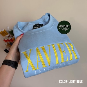 May include: A light blue crewneck sweatshirt featuring the word XAVIER embroidered in large yellow capital letters, with the word INSTITUTE embroidered in smaller white capital letters underneath. The garment has a black interior neck label.