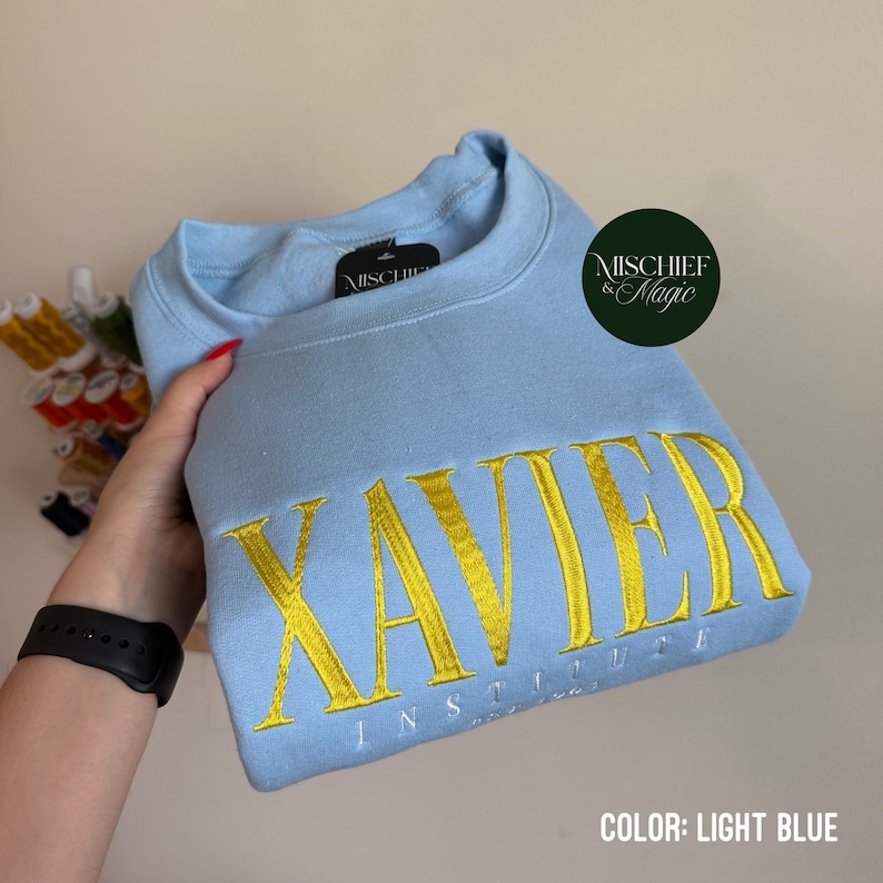May include: A light blue crewneck sweatshirt featuring the word XAVIER embroidered in large yellow capital letters, with the word INSTITUTE embroidered in smaller white capital letters underneath. The garment has a black interior neck label.