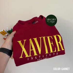 May include: A garnet-coloured crewneck sweatshirt featuring the embroidered yellow text XAVIER INSTITUTE EST 1964 across the chest. The garment has a round neckline and long sleeves, with a circular logo patch in the upper right corner reading MISCHIEF & MAGIC.