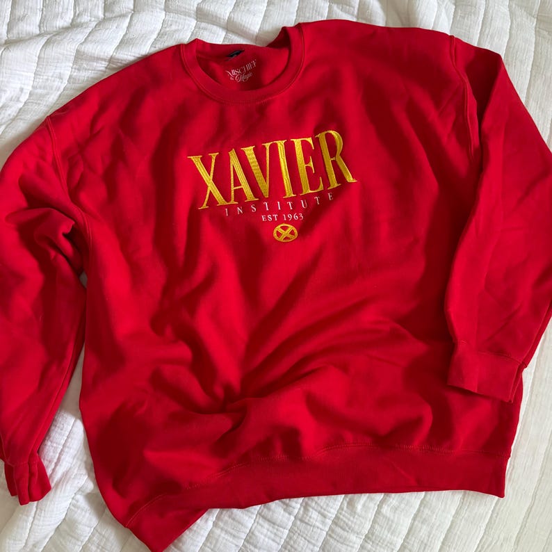 May include: A bright red crewneck sweatshirt featuring yellow embroidered text that reads XAVIER INSTITUTE EST 1963 with a circular X logo underneath. The garment has long sleeves and a ribbed collar, cuffs, and hem, showcasing a bold design.