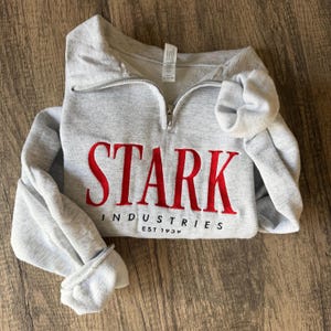 Könnte beinhalten: Ein hellgraues Sweatshirt mit kurzem Reißverschluss, auf dessen Vorderseite der Schriftzug STARK in roter Stickerei und INDUSTRIES EST 1939 in schwarzer Stickerei zu sehen ist. Das Kleidungsstück hat lange Ärmel, einen silbernen Reißverschluss und gerippte Bündchen.