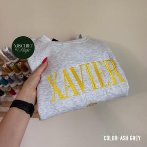 May include: An ash grey crewneck sweatshirt featuring the name XAVIER embroidered in bold yellow capital letters across the chest. The fabric has a heathered texture, and the garment is shown with a dark green circular logo tag attached.