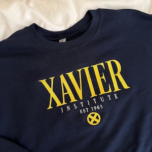 May include: A navy blue crewneck sweatshirt featuring yellow embroidered text that reads XAVIER INSTITUTE EST 1963. Below the text is a yellow circular emblem with an X symbol inside. The garment has long sleeves and a ribbed collar.
