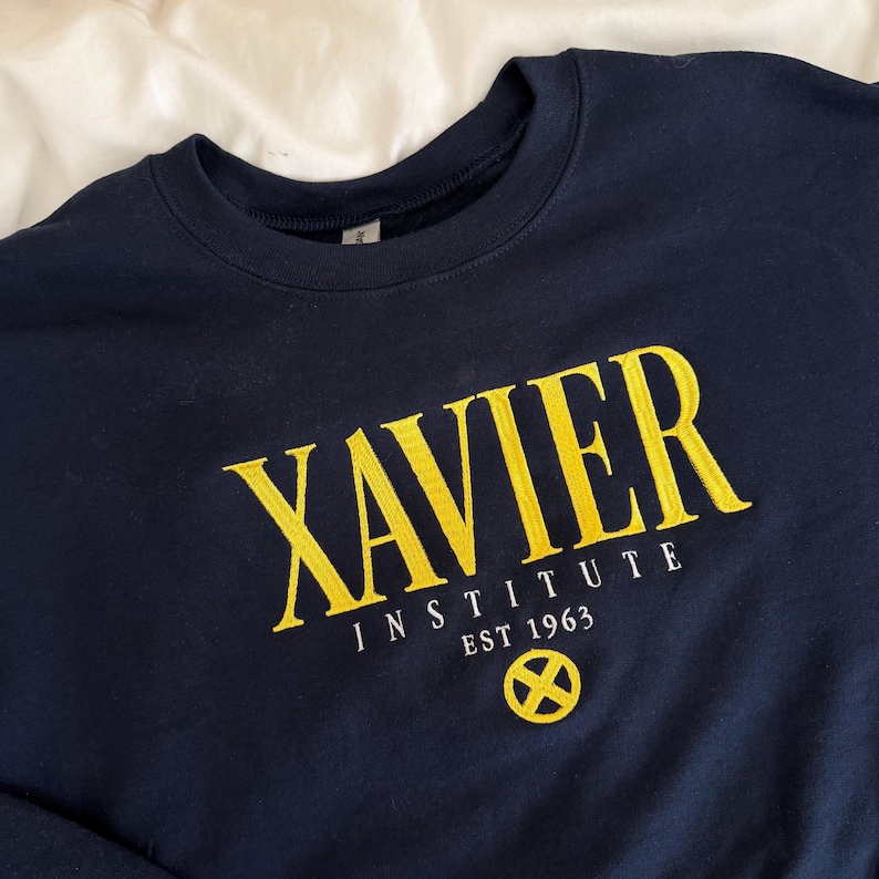 May include: A navy blue crewneck sweatshirt featuring yellow embroidered text that reads XAVIER INSTITUTE EST 1963. Below the text is a yellow circular emblem with an X symbol inside. The garment has long sleeves and a ribbed collar.