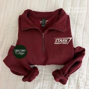 May include: A maroon quarter-zip pullover sweatshirt featuring a white embroidered Stark Industries logo on the chest. The garment has a high collar with a matching maroon zipper pull and long sleeves with ribbed cuffs. A black brand tag is at the neck.
