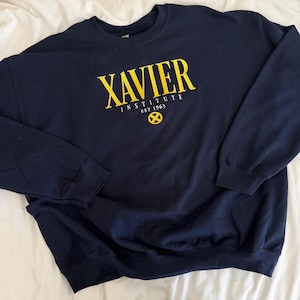 May include: A navy blue crewneck sweatshirt featuring yellow screen-printed text that reads XAVIER INSTITUTE EST 1963, accompanied by a yellow circular X-Men logo emblem positioned directly below the text on the centre of the chest.