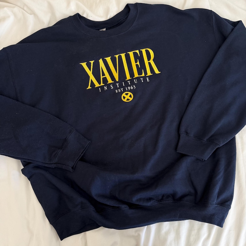 May include: A navy blue crewneck sweatshirt featuring yellow screen-printed text that reads XAVIER INSTITUTE EST 1963, accompanied by a yellow circular X-Men logo emblem positioned directly below the text on the centre of the chest.