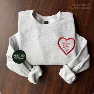 Könnte beinhalten: Ein hellgrau meliertes Sweatshirt mit Rundhalsausschnitt und einem roten gestickten Herz auf der linken Brust. Im Herz steht der Text TO GROW OLD IN V. Ein kreisförmiges dunkelgrünes Logo mit dem Text MISCHIEF & MAGIC ist links zu sehen.