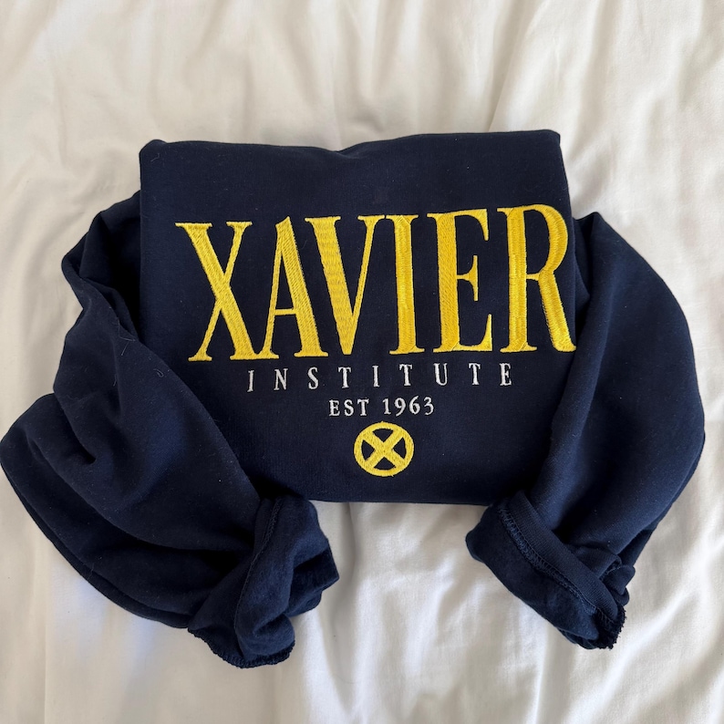 May include: A navy blue long-sleeve sweatshirt featuring the yellow embroidered text XAVIER INSTITUTE, with the white text EST 1963 and a yellow circular X logo centred below. The garment has a crew neckline and long sleeves with ribbed cuffs.