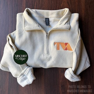 Può includere: Una felpa color crema con zip a un quarto, colletto alto e maniche lunghe. Sul petto è presente un logo ricamato arancione e rosso con le lettere TVA. È visibile un logo circolare verde scuro con il testo Mischief & Magic.