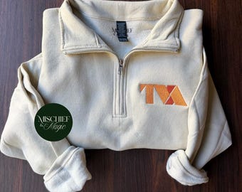 May include: A cream-colored quarter-zip sweatshirt featuring a stand-up collar and long sleeves. The chest displays an embroidered orange and red logo with the letters TVA. A dark green circular logo with the text Mischief & Magic is present.