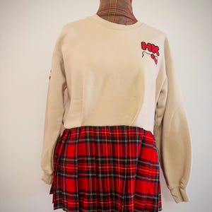 Könnte beinhalten: Ein beiges Langarm-Sweatshirt mit dem roten Schriftzug HK und einer kleinen roten Schleifengrafik auf der Brust. Der untere Teil des Kleidungsstücks ist mit einem roten, schwarzen und grünen Faltenrock mit klassischem Schottenkaro verbunden.